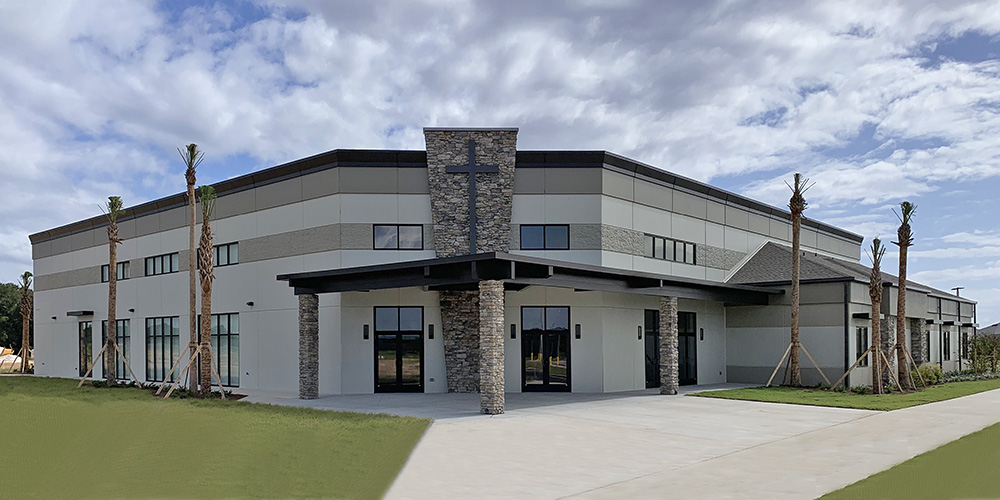 Village Park Church - a light and dark gray metal building project with blue skies and clouds behind it and green grass and a sidewalk in front.