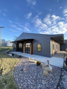 the front of a beautiful, gray steel building with three large windows and blue skies behind it with a sidewalk in front and around the side.