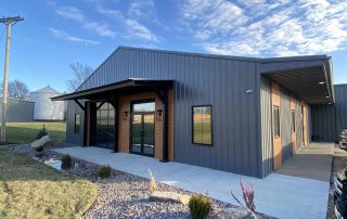 The front of a beautiful, gray pre-engineered metal building with three large windows and blue skies behind it with a sidewalk in front and around the side.