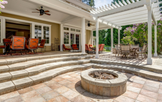 A large, off-white patio design with a fire pit and concrete stairs along with red furniture and a white awning.